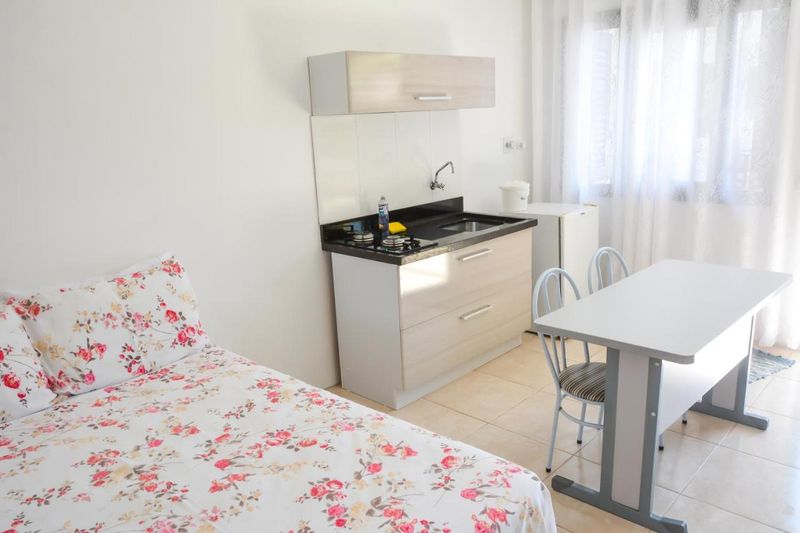 Quarto de casal com roupa de cama floral, cozinha compacta e mesa com cadeiras