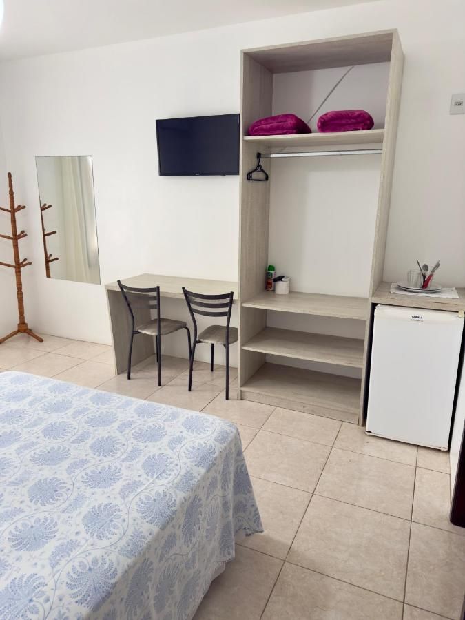 Quarto com TV de tela plana, armário aberto, mesa com cadeiras e frigobar